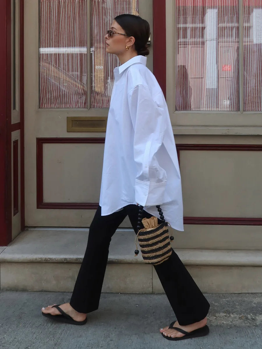 Woman walking outdoors wearing a white shirt and black pants with a striped bag.