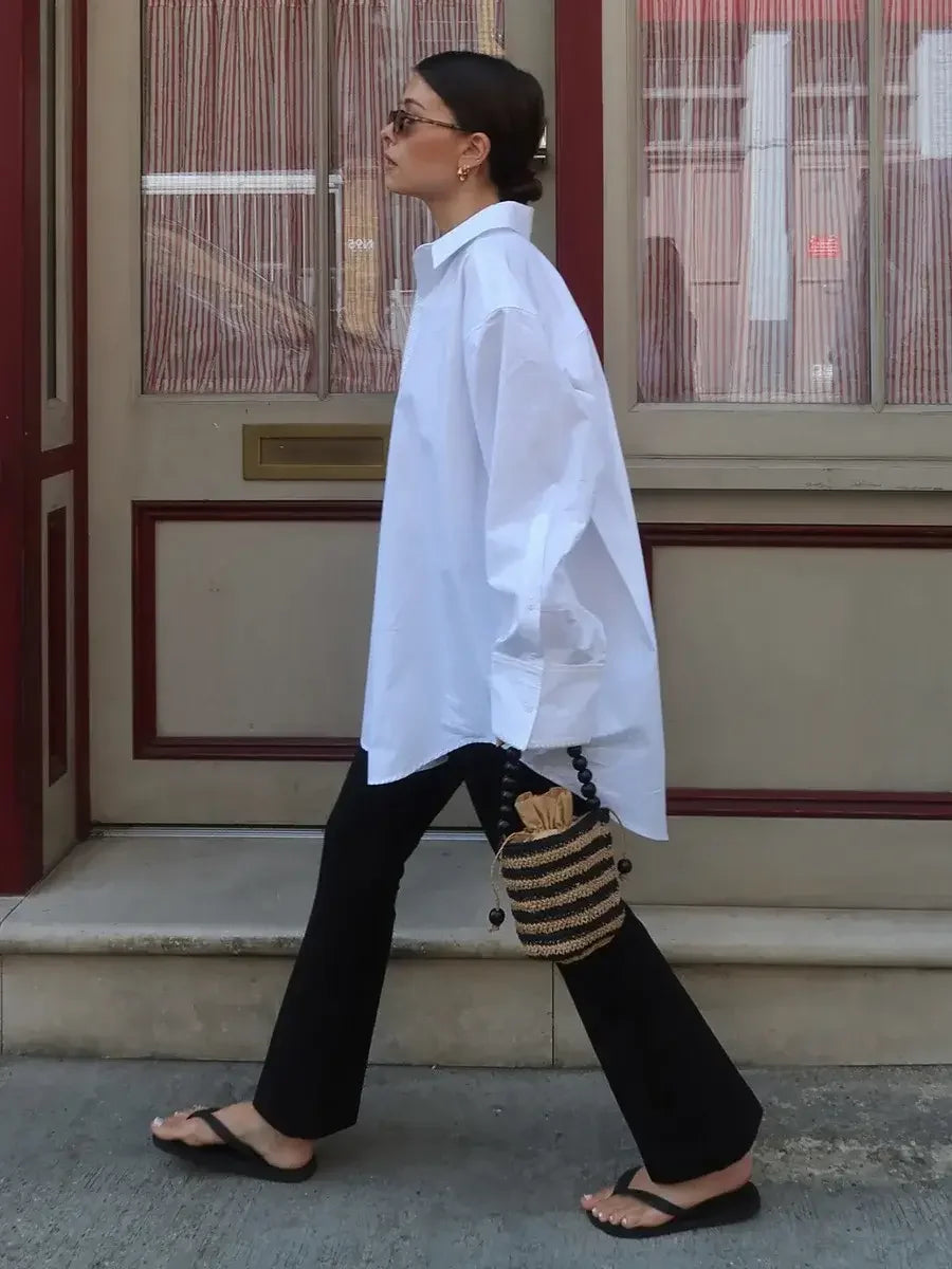 Woman walking outdoors wearing a white shirt and black pants with a striped bag.