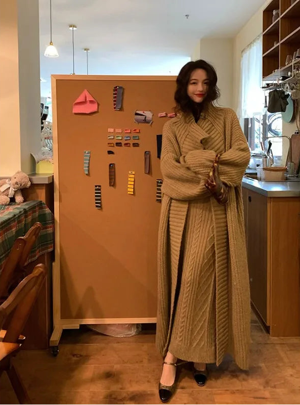 Woman in a long beige coat standing in a kitchen with a refrigerator covered by a brown paper bag.