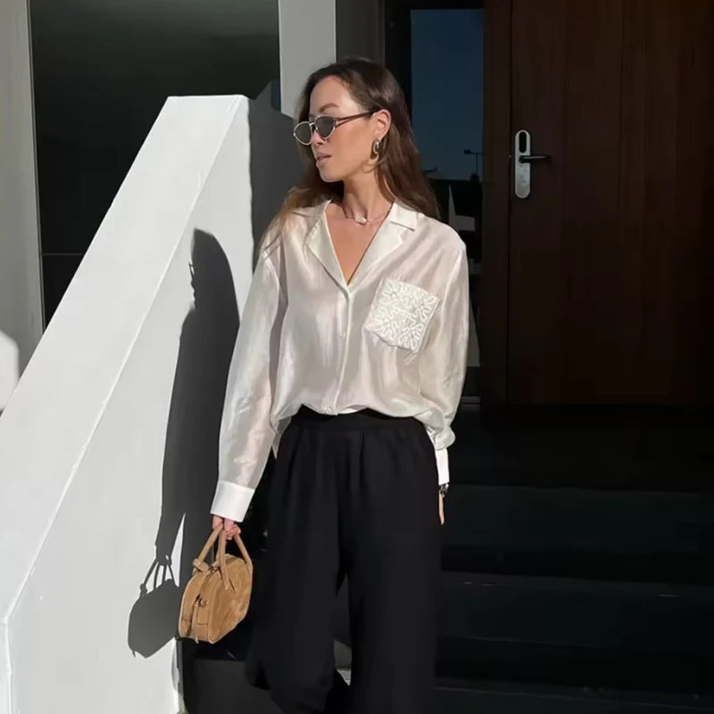 Woman in a white blouse with a pocket and black pants holding a beige handbag, standing in front of a building.