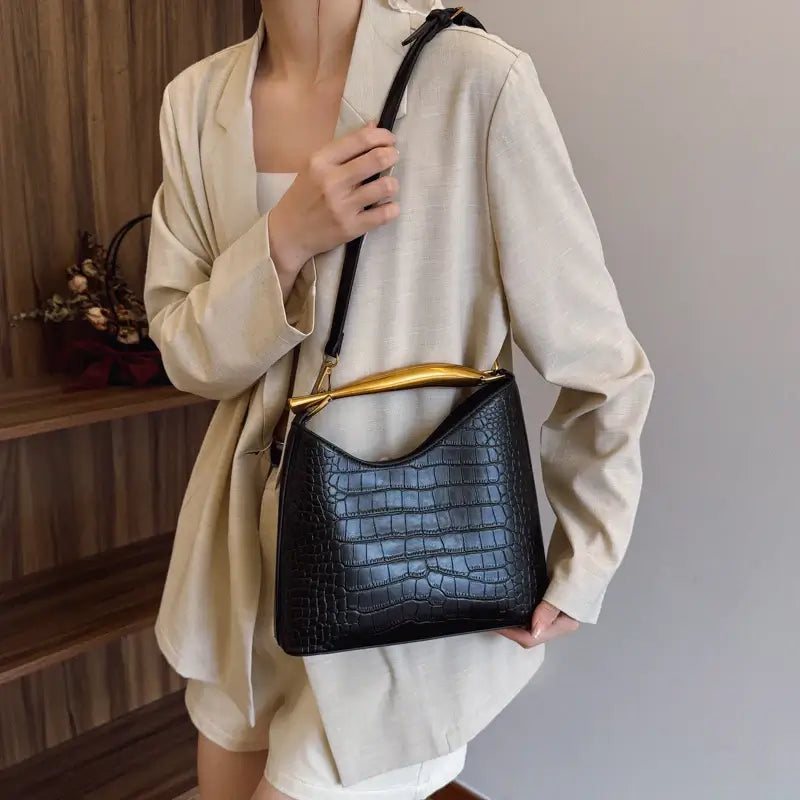 Person holding a black crocodile-patterned handbag with a gold handle.