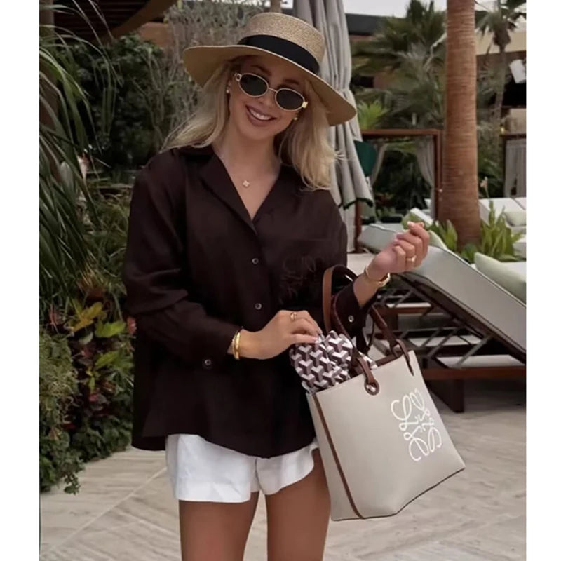 Woman in a brown shirt and white shorts holding a beige tote bag with a logo, standing outdoors.