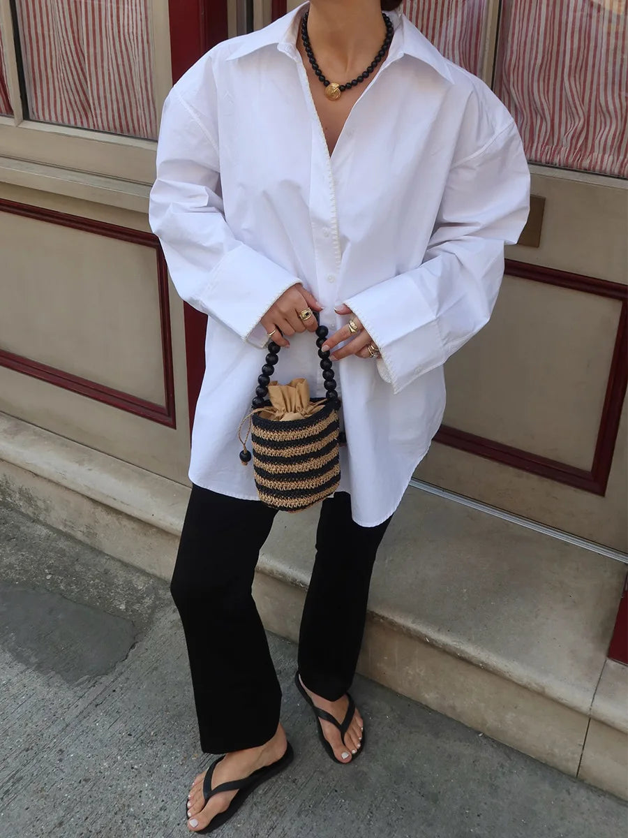 Person wearing a white shirt and black pants holding a woven bag on a sidewalk.