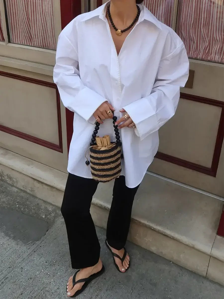 Person wearing a white shirt and black pants holding a woven bag on a sidewalk.