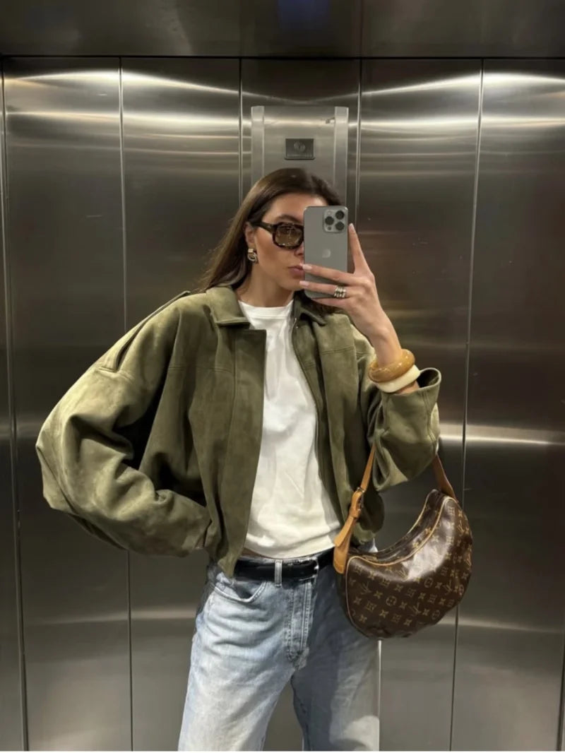 Person taking a mirror selfie in an elevator wearing a green jacket, white shirt, and blue jeans.