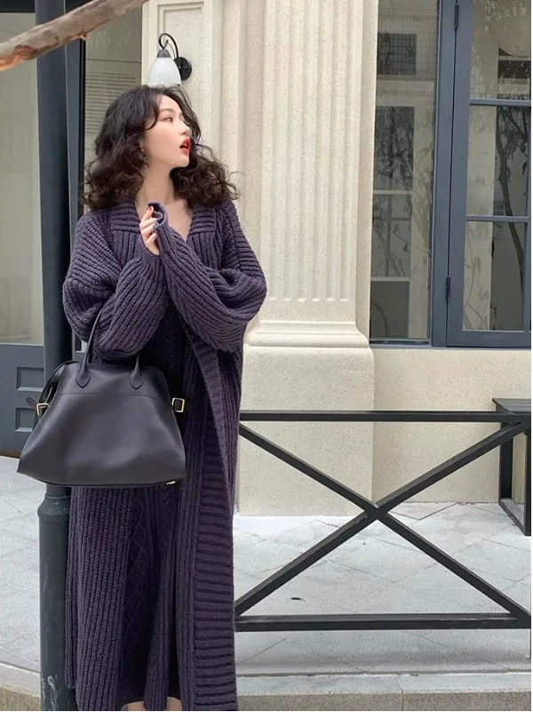 Woman in a long dark purple cardigan holding a black handbag outdoors.