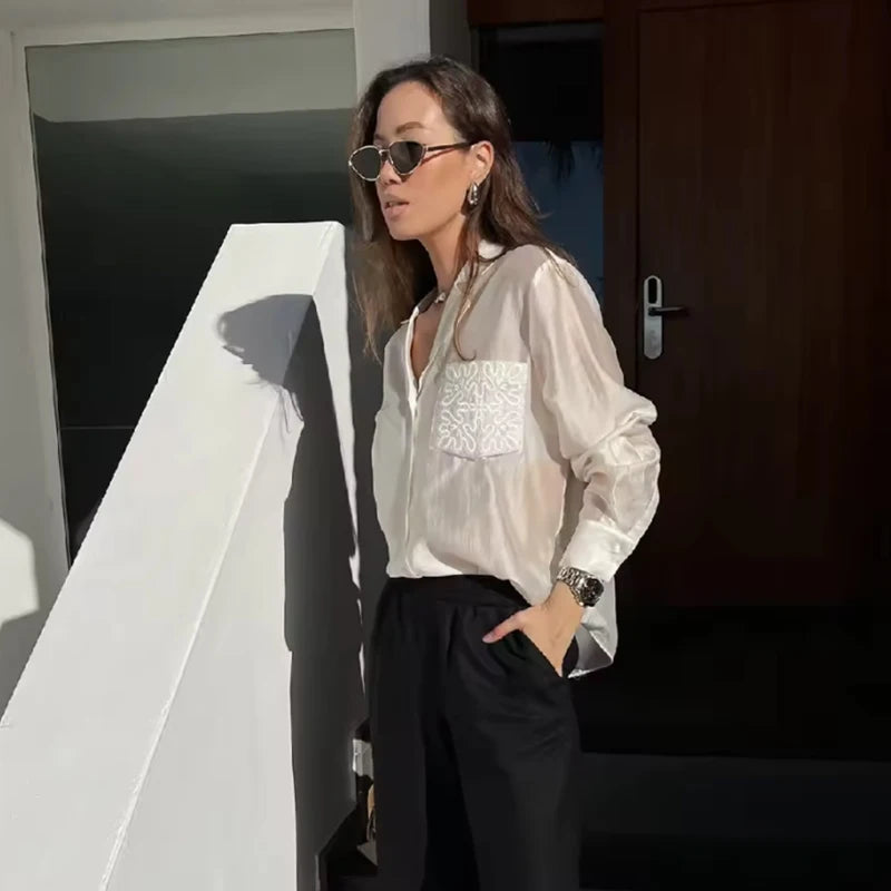 Woman wearing a white blouse with lace details and black pants, standing against a dark wall.