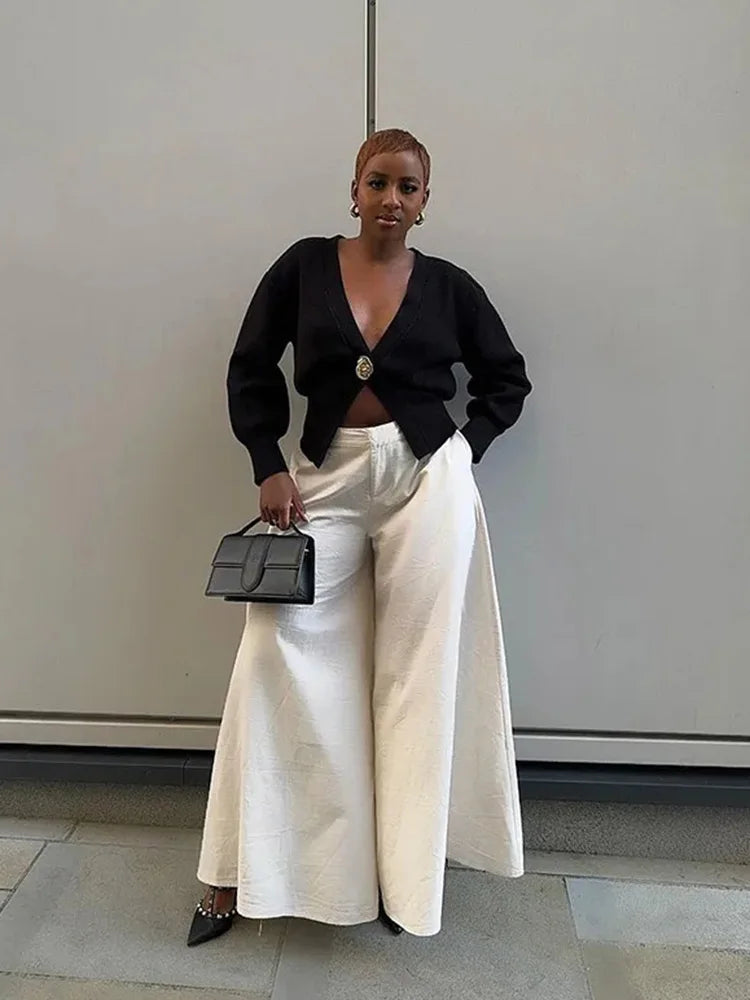 Woman in black top and white pants holding a black handbag against a plain background