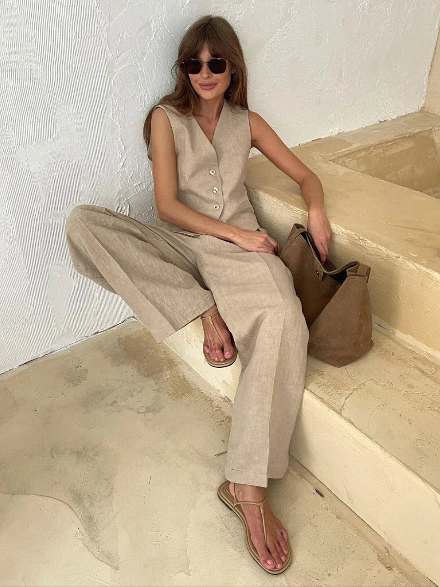 Woman in beige sleeveless jumpsuit sitting on a stone step holding a brown bag.