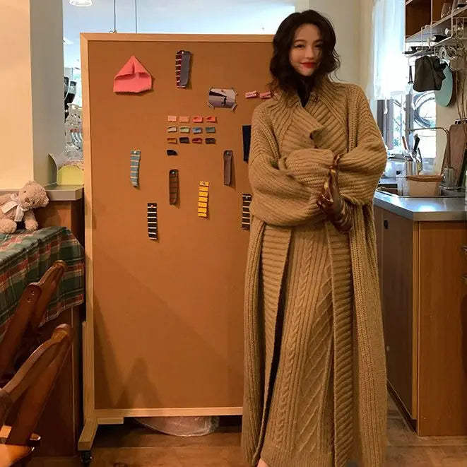 Woman in a long beige cardigan standing in a kitchen with a wooden board behind her.