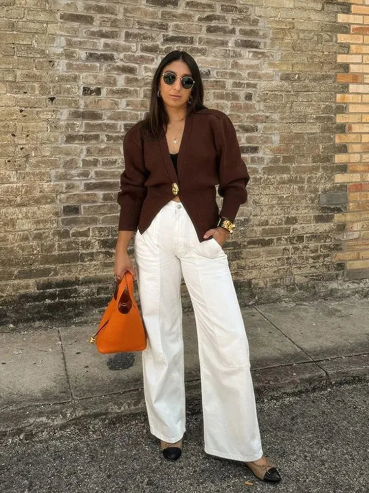 Woman in brown jacket and white pants standing against a brick wall.