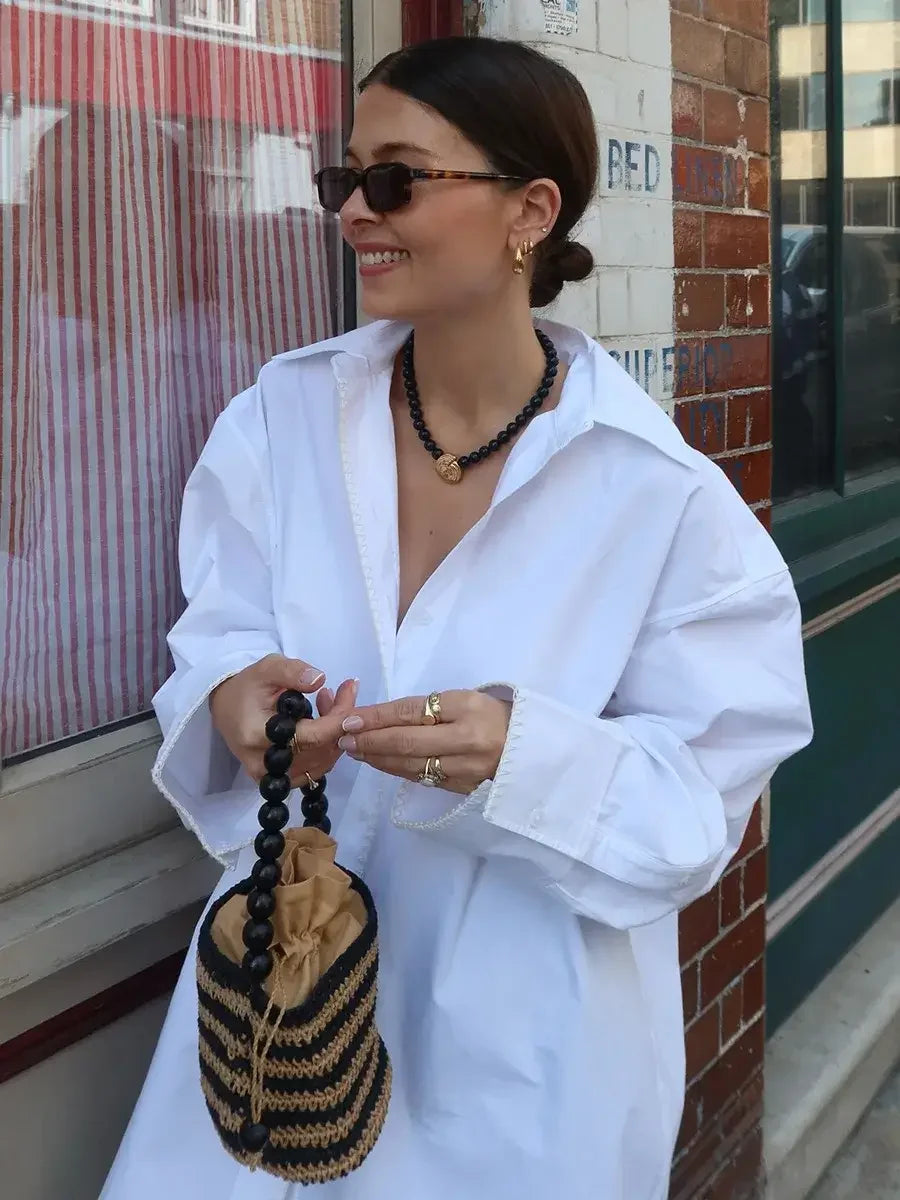Woman in a white shirt holding a black and beige handbag, standing in front of a brick wall.
