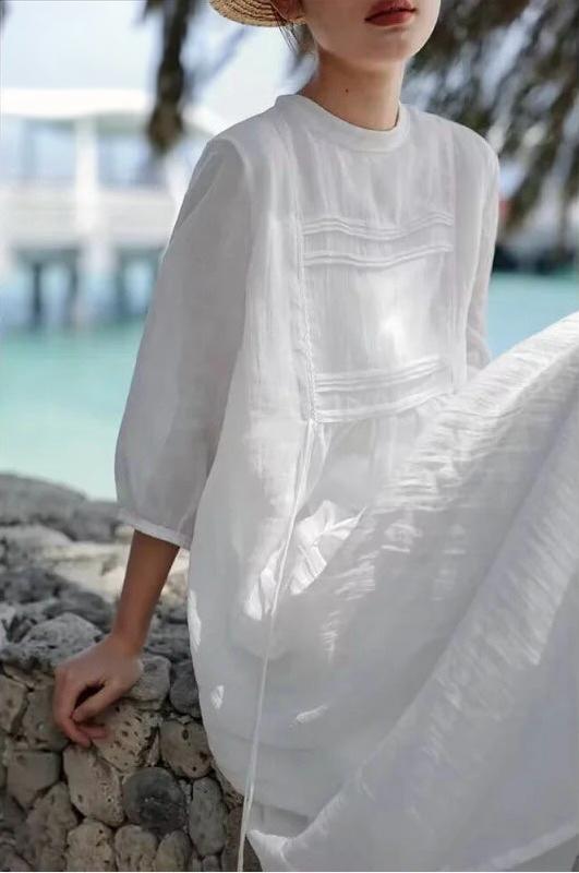 Side view of woman seated in a loose-fit white pintuck maxi dress holding a woven straw tote