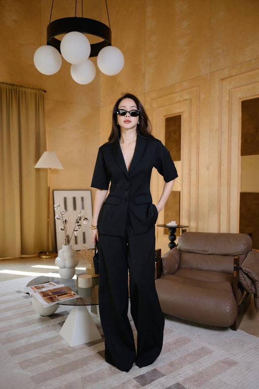A woman confidently walking outdoors wearing the black 2-piece tailored suit set, featuring a short-sleeve blazer with a deep V-neck and high-waisted wide-leg trousers, styled with sunglasses and a small handbag.
