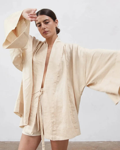 A woman modeling a light beige linen kimono robe with wide bell sleeves and a matching tie belt, set against a neutral light gray wall.