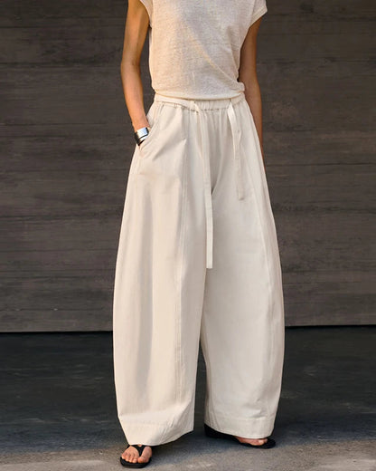 A person wearing loose, wide-leg white pants with a drawstring waist and side pockets, paired with a light sleeveless top and black sandals, standing against a dark wood-paneled wall.