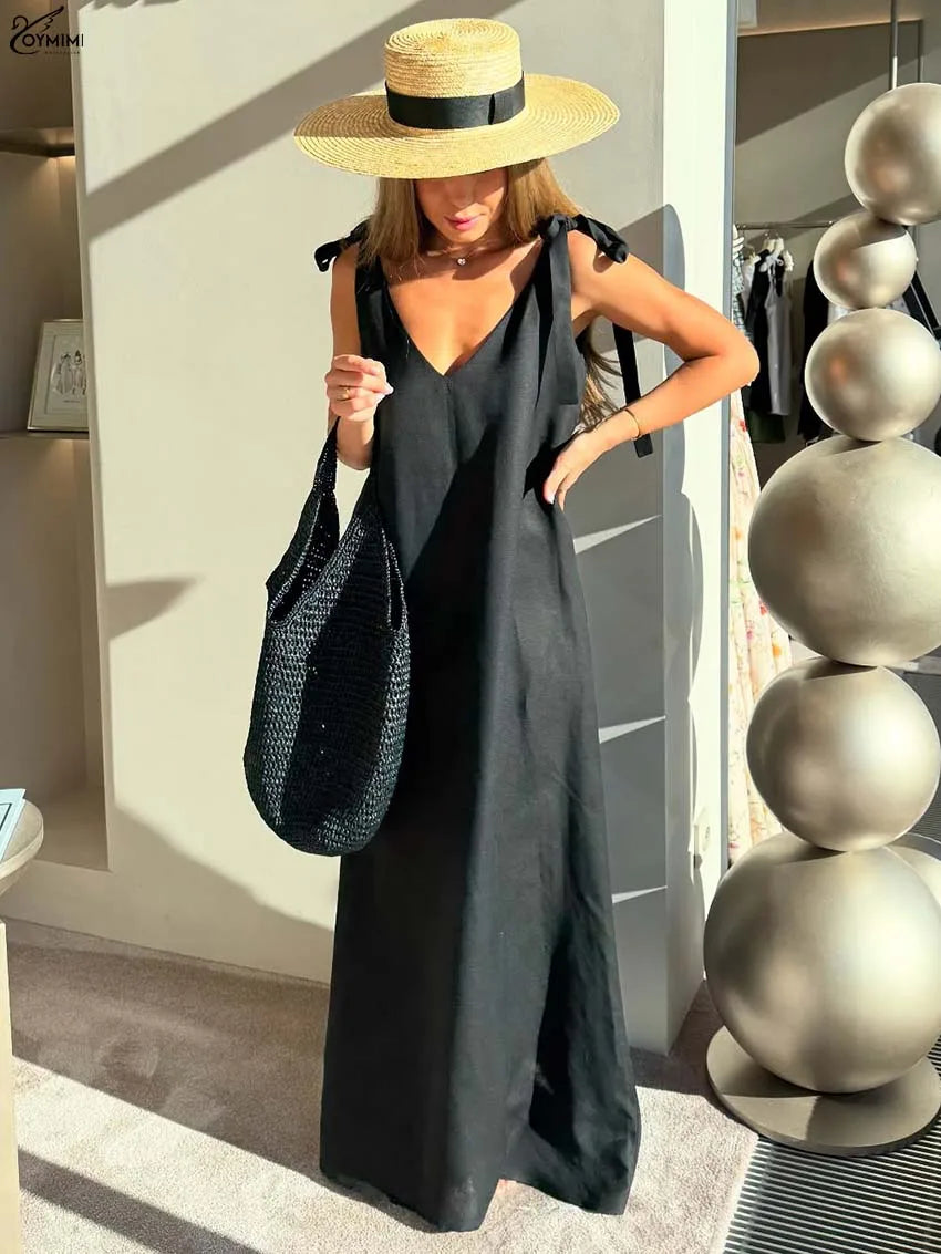 A woman wearing a black sleeveless maxi dress with tie shoulder straps, a wide-brimmed straw hat with a black band, and carrying a black woven tote bag, standing in a modern indoor setting with a decorative metallic sphere sculpture.