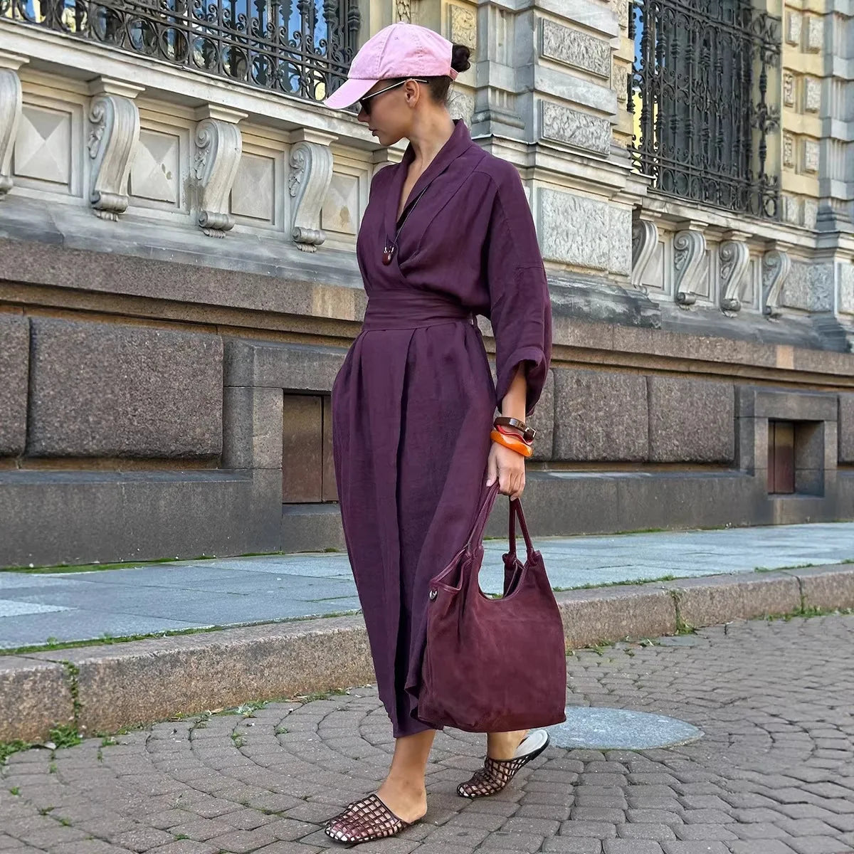 A woman wearing a dark purple midi wrap dress, a pink baseball cap, and patterned flat shoes walks on a cobblestone street, holding a dark purple suede tote bag.