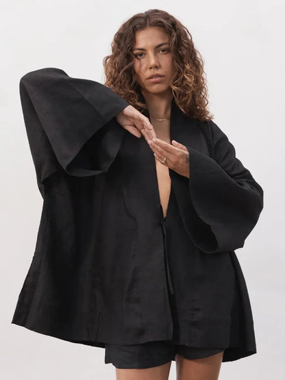 A model wearing a black satin-like long-sleeve jacket with dramatic bell sleeves and matching black shorts, posed against a light gray studio background.