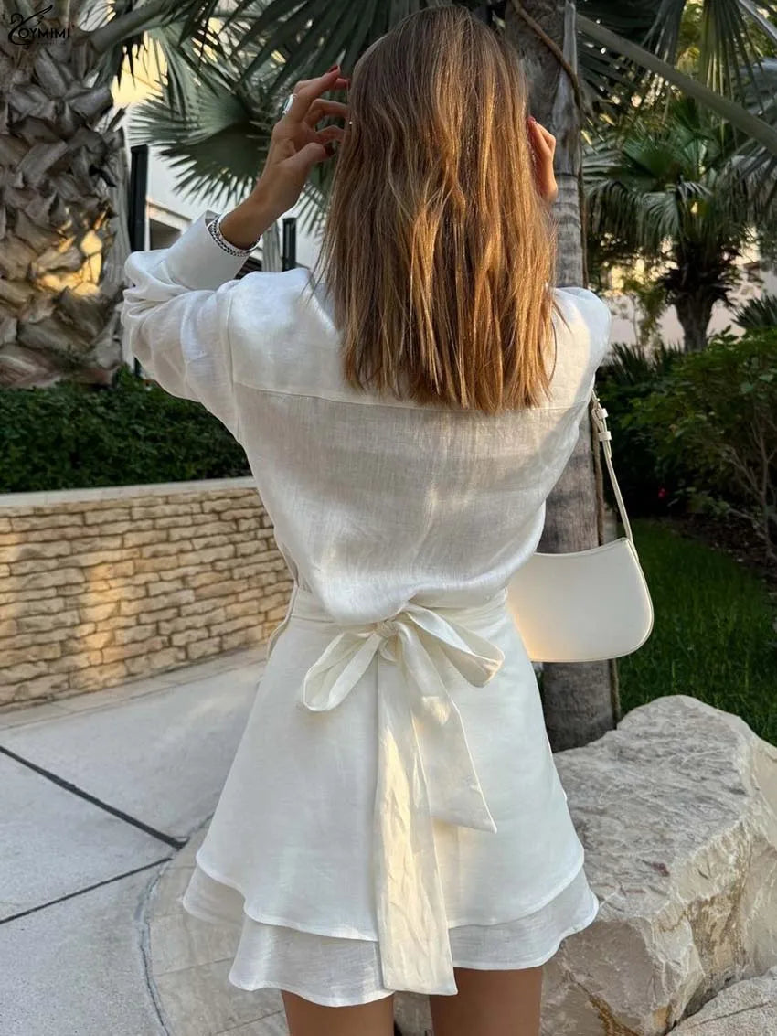 Woman in a white outfit standing outdoors with plants and stone wall in the background