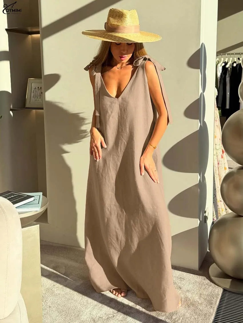 A woman wearing a sleeveless taupe maxi dress and a wide-brimmed straw hat, standing in a modern indoor setting with strong sunlight and shadows.