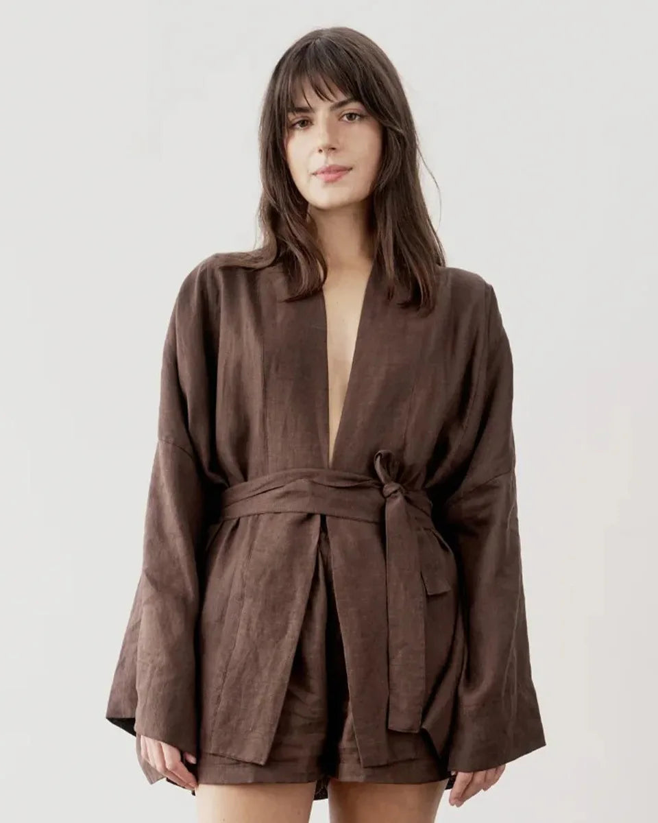 A woman modeling a brown linen robe with a tie waist belt, photographed against a plain light gray studio background.