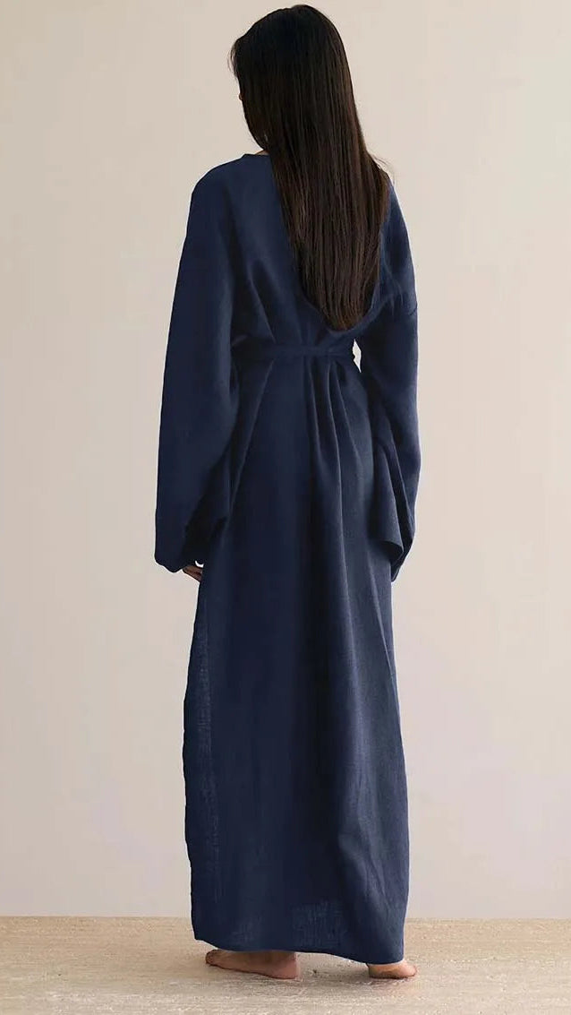 A woman viewed from behind wearing a long navy blue wrap robe or kimono-style dress with a tie at the back, standing barefoot against a plain light beige wall.