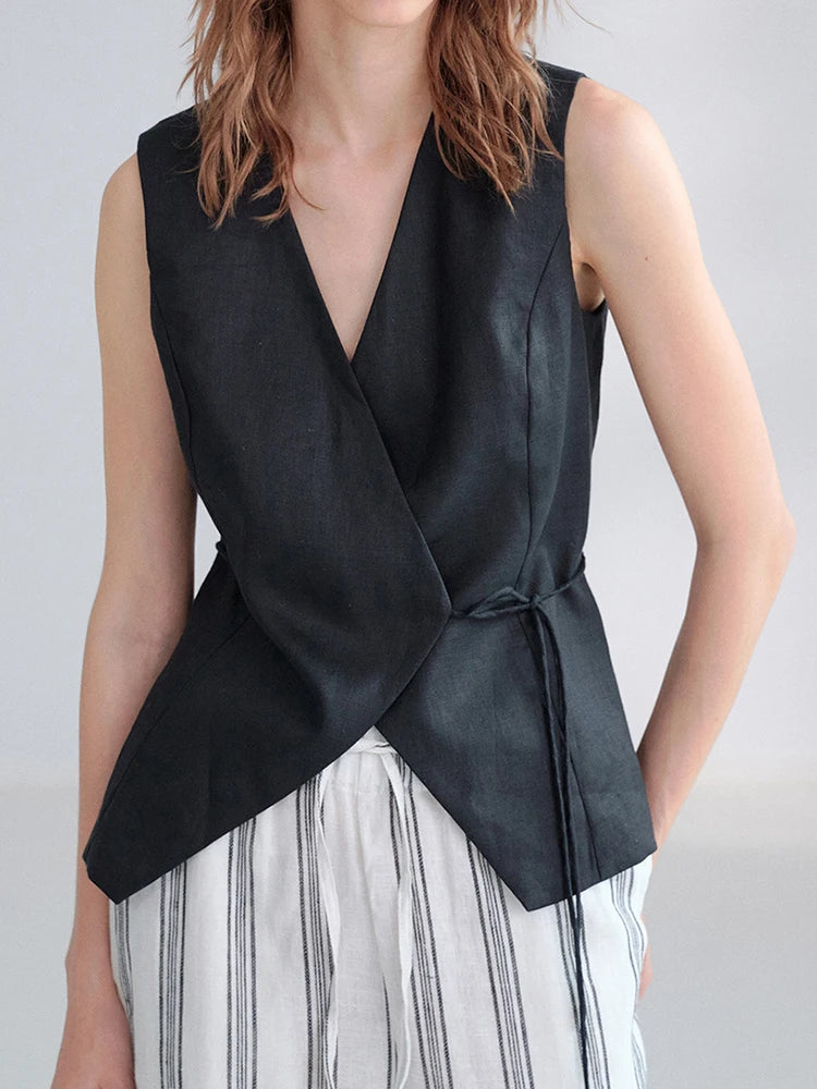 A sleeveless black wrap-style vest with a deep V-neck and self-tie waist, worn by a model over white and gray striped trousers against a light gray studio background.