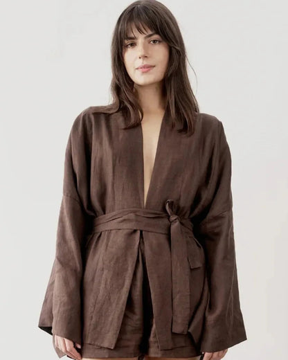 A woman wearing a dark brown linen wrap robe with a tie waist belt, photographed against a plain light gray studio background.