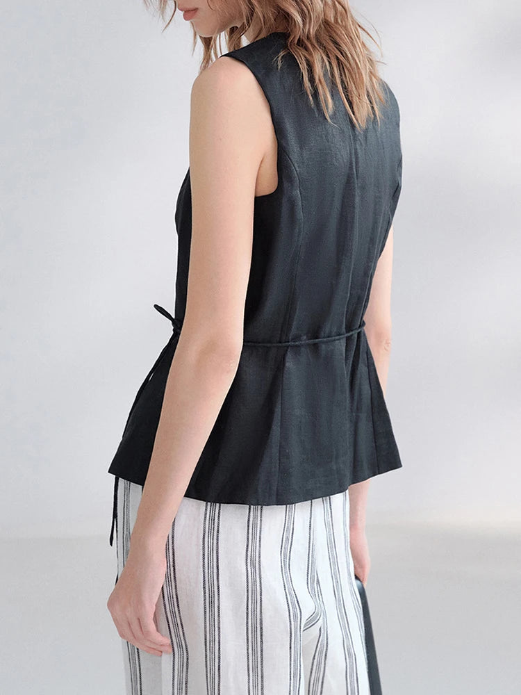 A woman shown from the back wearing a sleeveless black peplum-style top with a tie detail at the waist and white pants with thin black vertical stripes, against a light gray studio background.
