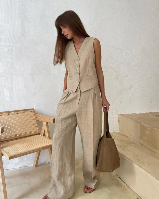 Woman wearing a beige linen outfit with wide-leg pants and a vest, standing in a minimalistic room.