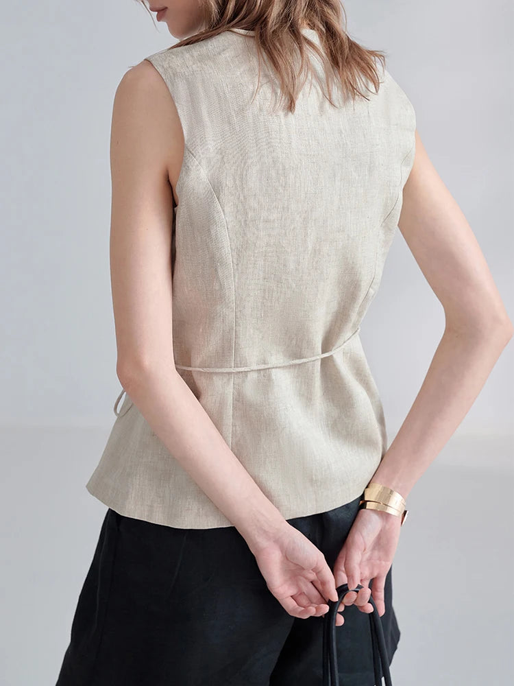 A woman shown from the back wearing a sleeveless light beige linen top with a tie waist detail and black trousers, set against a plain light gray studio background.