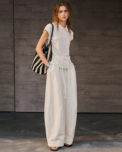 A woman stands against a dark wood-paneled wall wearing a light beige sleeveless collared top and matching wide-leg pants, with a black-and-white striped shoulder bag.