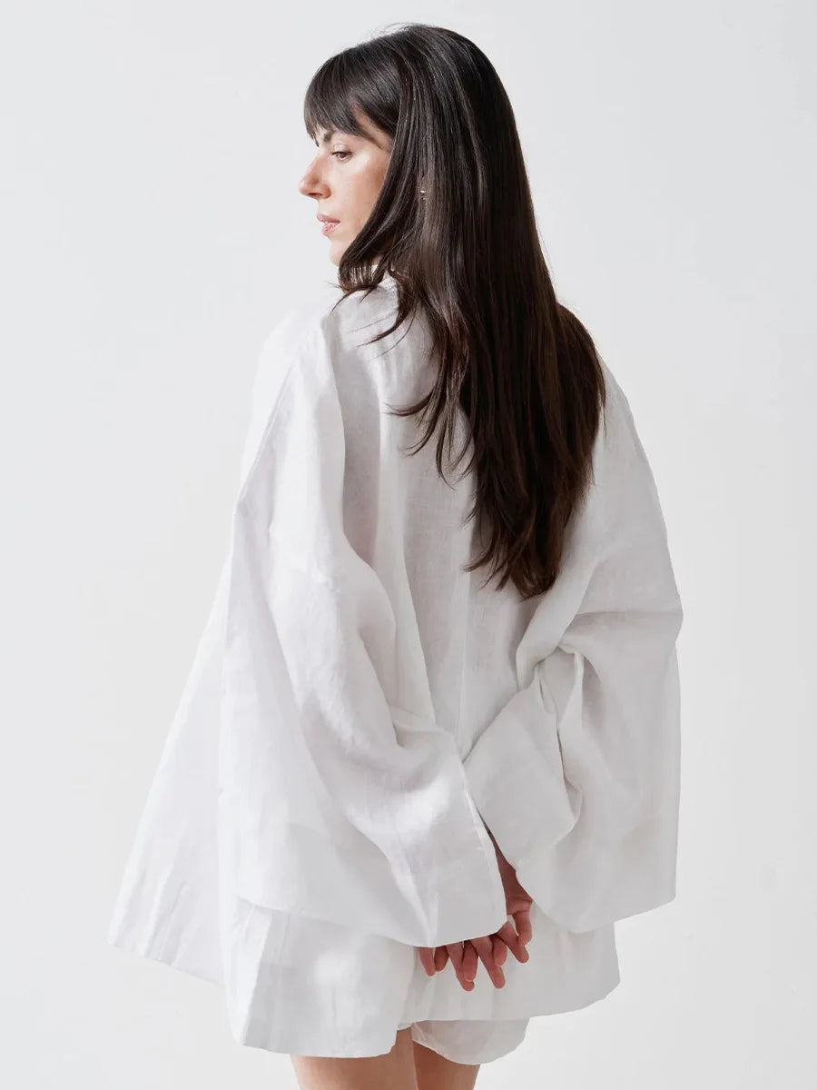 A woman with long dark hair is shown from the back wearing a loose, oversized white linen shirt with voluminous sleeves, set against a plain light gray studio background.