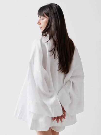 A woman with long dark hair is shown from the back wearing a loose, oversized white linen shirt with voluminous sleeves, set against a plain light gray studio background.