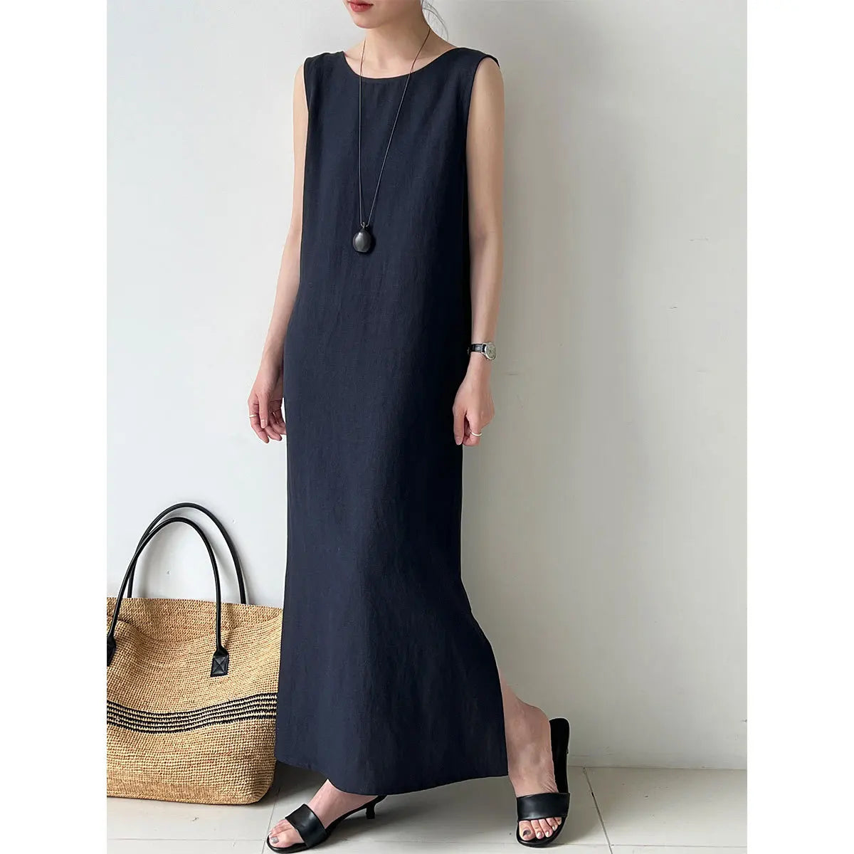 A woman wearing a sleeveless, dark navy maxi dress with a relaxed fit, standing next to a woven straw tote bag. She is wearing black slide sandals and a long pendant necklace.