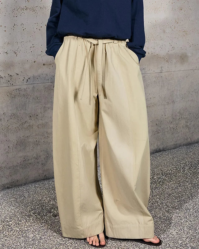 A person wearing loose, wide-leg beige drawstring pants with a navy long-sleeve top, standing barefoot on a speckled concrete floor against a textured concrete wall.