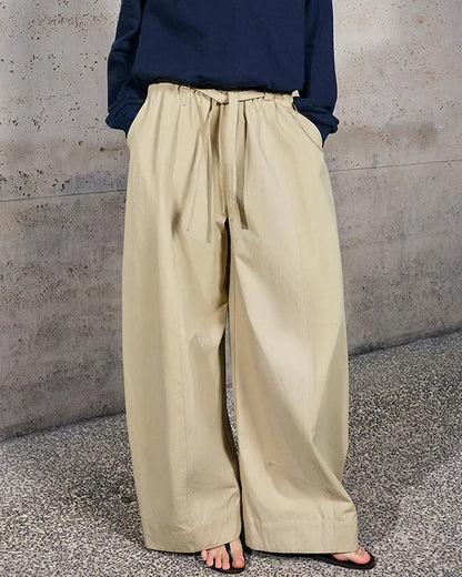 A person wearing loose, wide-leg beige drawstring pants with a navy long-sleeve top, standing barefoot on a speckled concrete floor against a textured concrete wall.