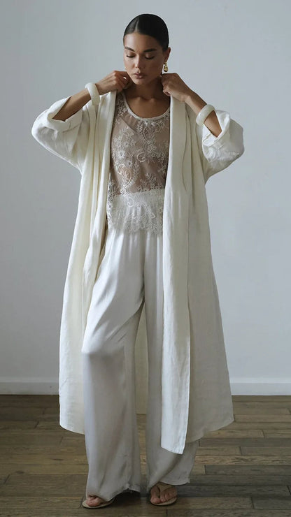A woman stands in a minimalist room with a light gray wall and wooden floor, wearing a long cream linen open-front cardigan over a white lace-trimmed top and matching wide-leg trousers.