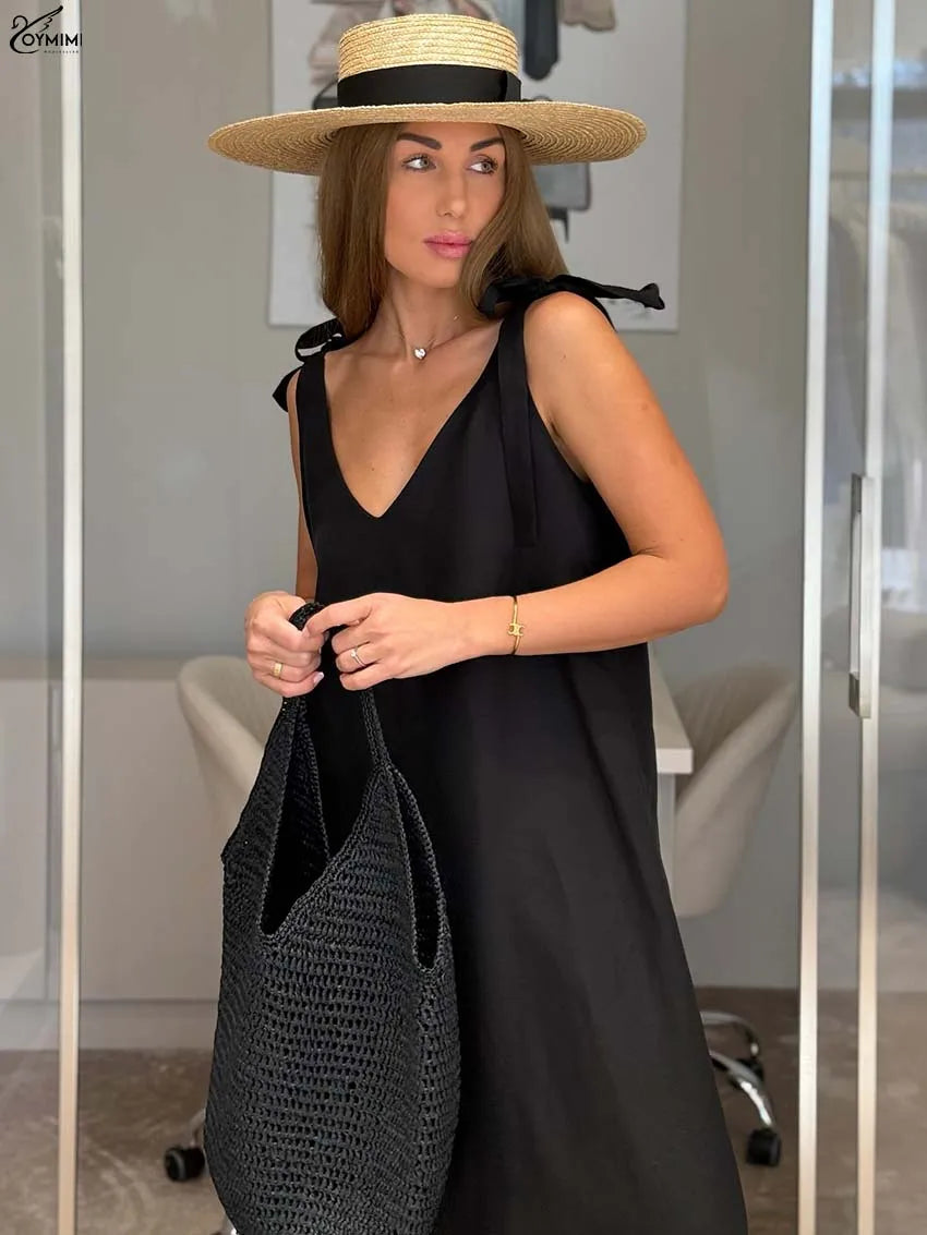 A woman wearing a black sleeveless V-neck dress, a wide-brimmed straw hat with a black ribbon, and carrying a large black woven tote bag, standing in front of a glass door.