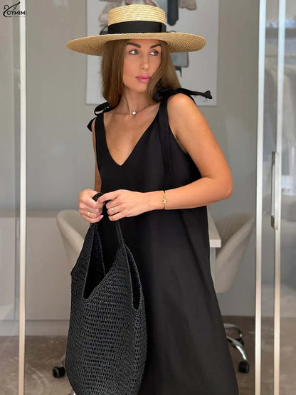 A woman wearing a black sleeveless V-neck dress, a wide-brimmed straw hat with a black ribbon, and carrying a large black woven tote bag, standing in front of a glass door.