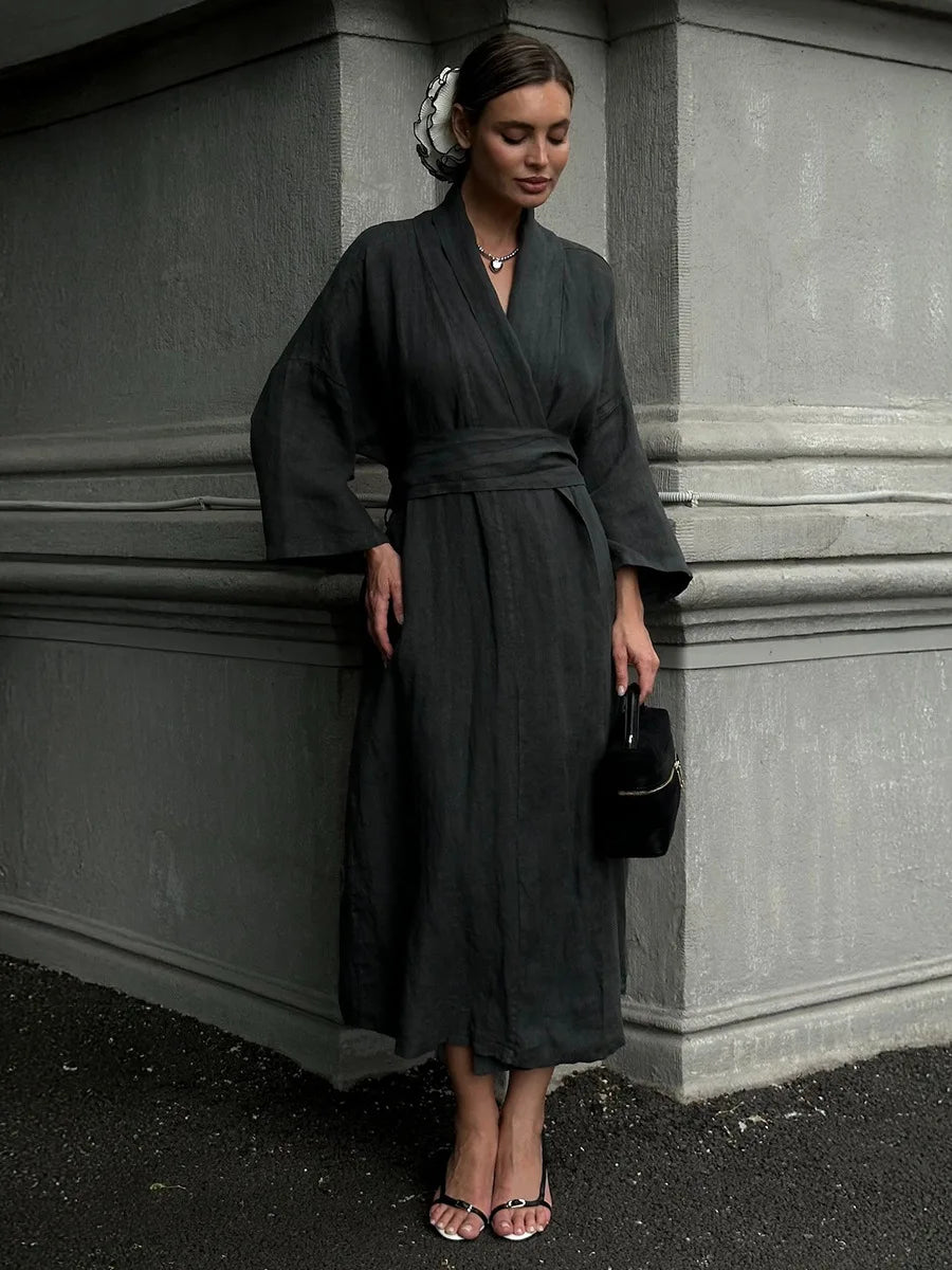 A woman wearing a dark charcoal gray wrap robe dress with wide sleeves and a matching waist tie, standing against a gray stone building corner.
