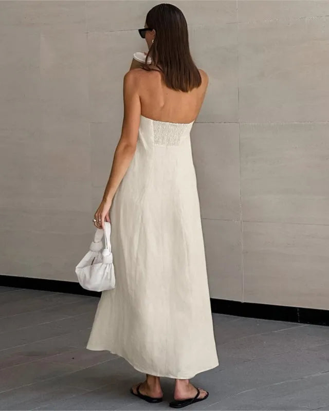 A woman wearing a strapless off-white maxi dress with a lace-up back, holding a white handbag and wearing black sandals, photographed from behind against a light gray tiled wall.