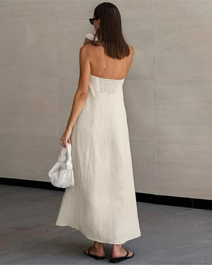 A woman wearing a strapless off-white maxi dress with a lace-up back, holding a white handbag and wearing black sandals, photographed from behind against a light gray tiled wall.