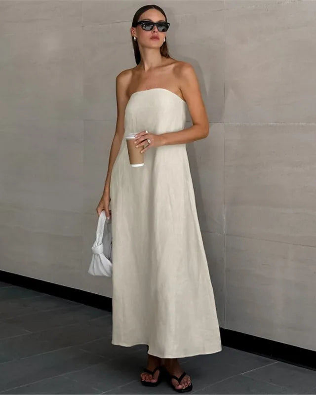 A woman stands against a light gray tiled wall wearing a strapless off-white maxi dress, holding a white bucket bag and a coffee cup.