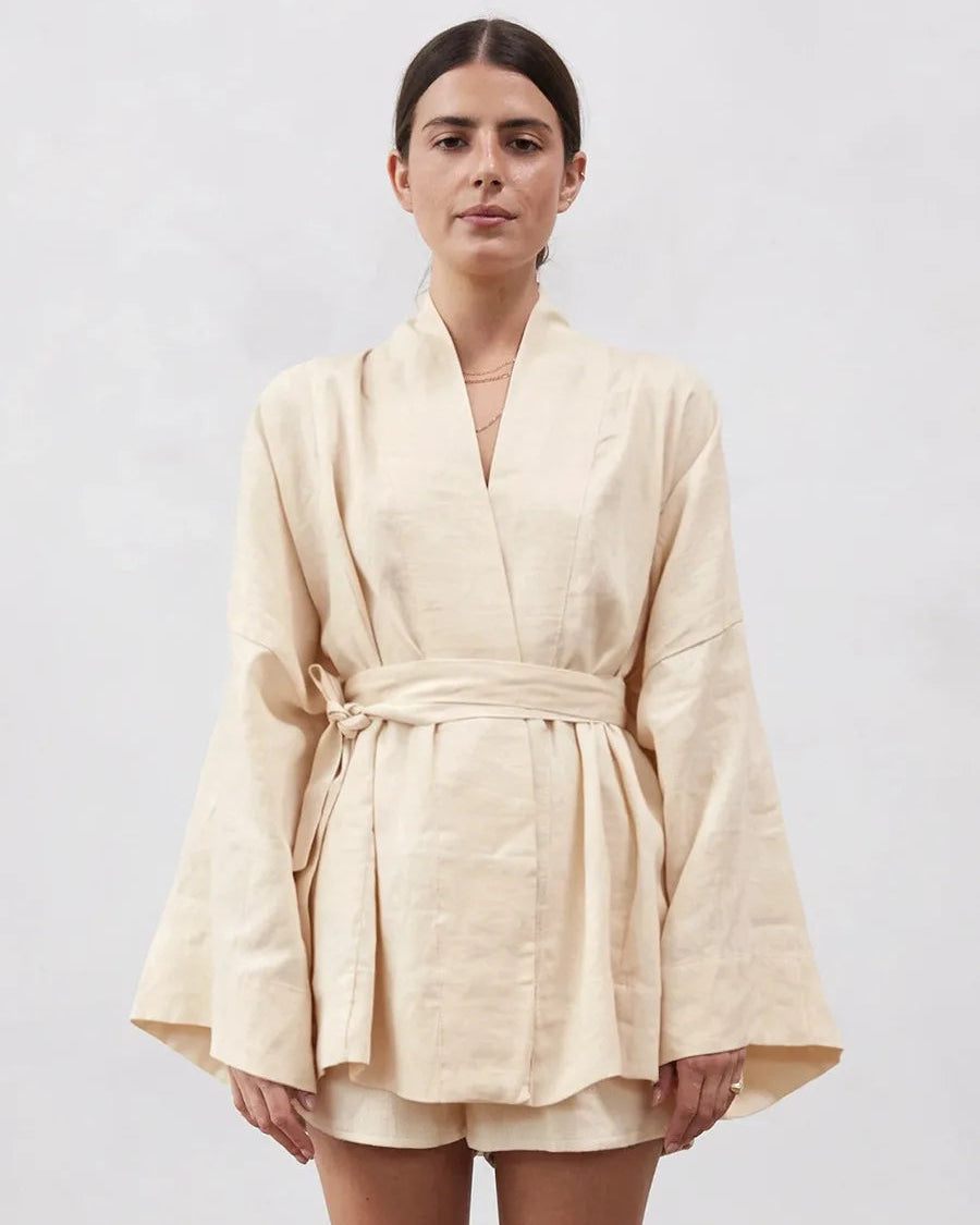 A woman modeling a light beige linen wrap robe with wide sleeves and a matching tie belt, paired with matching shorts, against a plain light gray background.