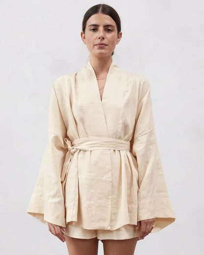 A woman modeling a light beige linen wrap robe with wide sleeves and a matching tie belt, paired with matching shorts, against a plain light gray background.