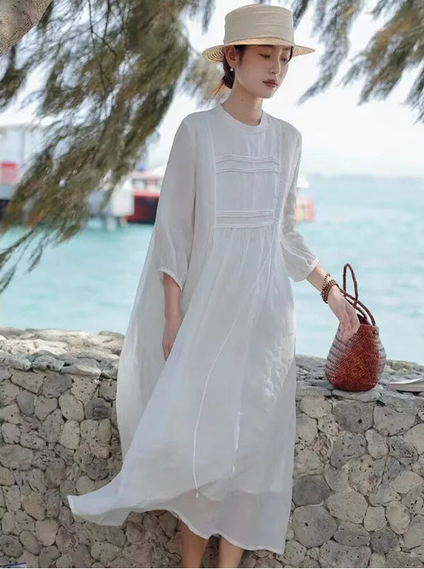 Woman wearing a white pintuck linen-blend maxi dress with 3/4 sleeves and straw hat by the seaside