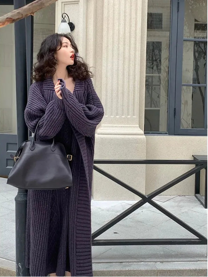 Woman in a long dark purple cardigan holding a black handbag outdoors.