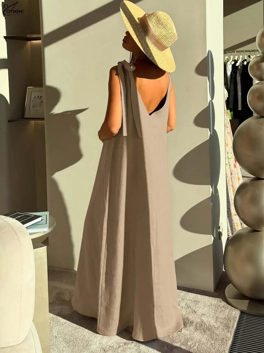 A woman stands with her back to the camera wearing a sleeveless taupe maxi dress with a tie-shoulder detail and a wide-brimmed straw hat in a sunlit boutique interior.